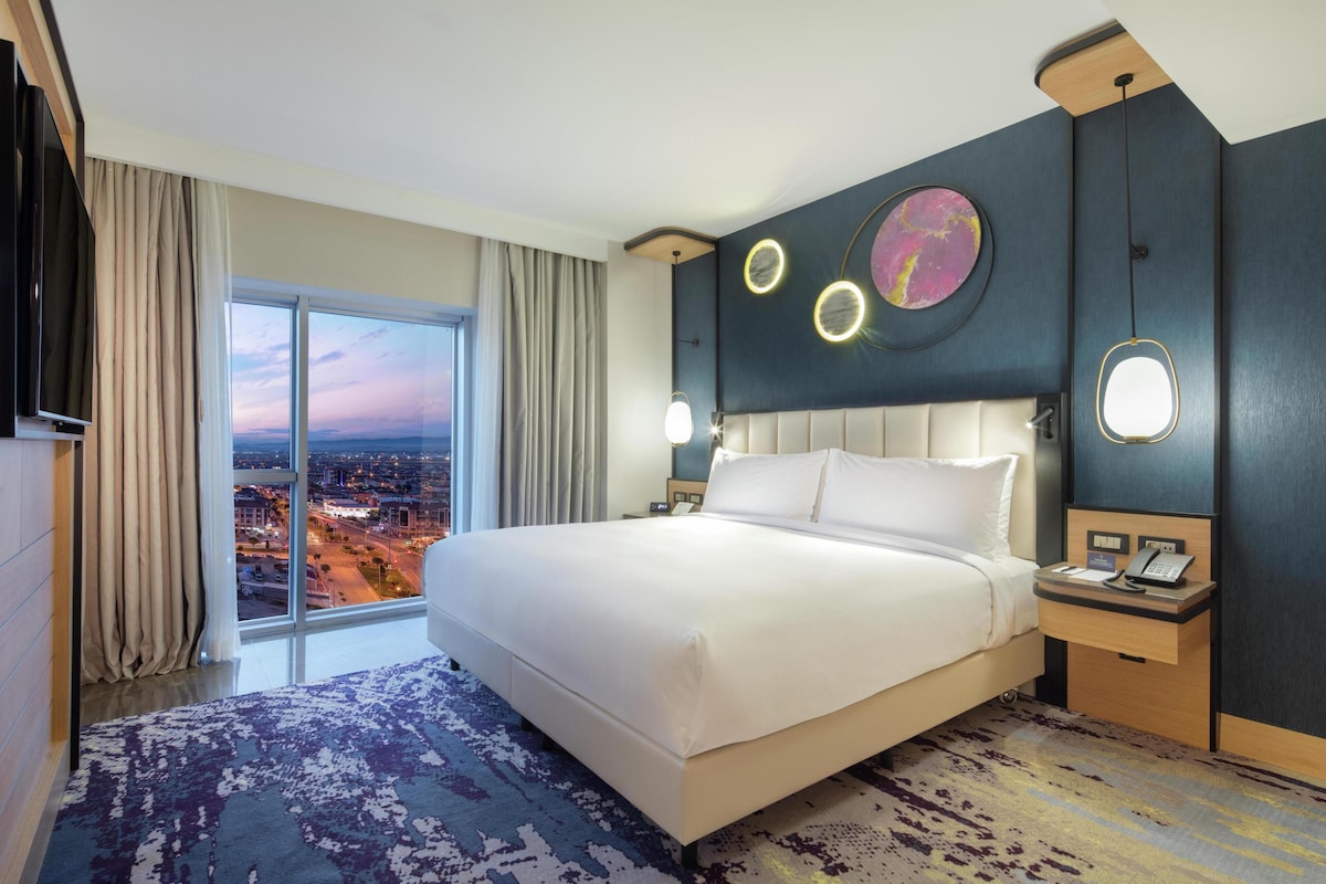 Habitación de hotel con una cama grande, televisión, mesita de noche con teléfono y vista a la ciudad a través de amplios ventanales.