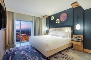 Habitación de hotel con una cama grande, televisión, mesita de noche con teléfono y vista a la ciudad a través de amplios ventanales.