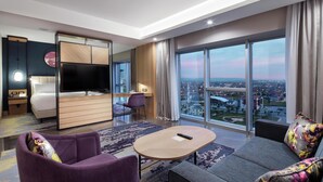 Una habitación de hotel moderna con una televisión grande, un sofá, una mesita y vistas a un paisaje urbano.