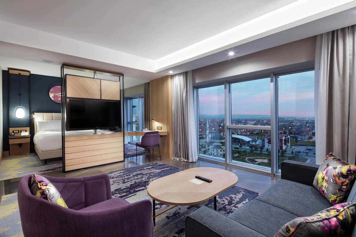A modern hotel room with a large TV, a sofa, a small table, and a view of a cityscape.