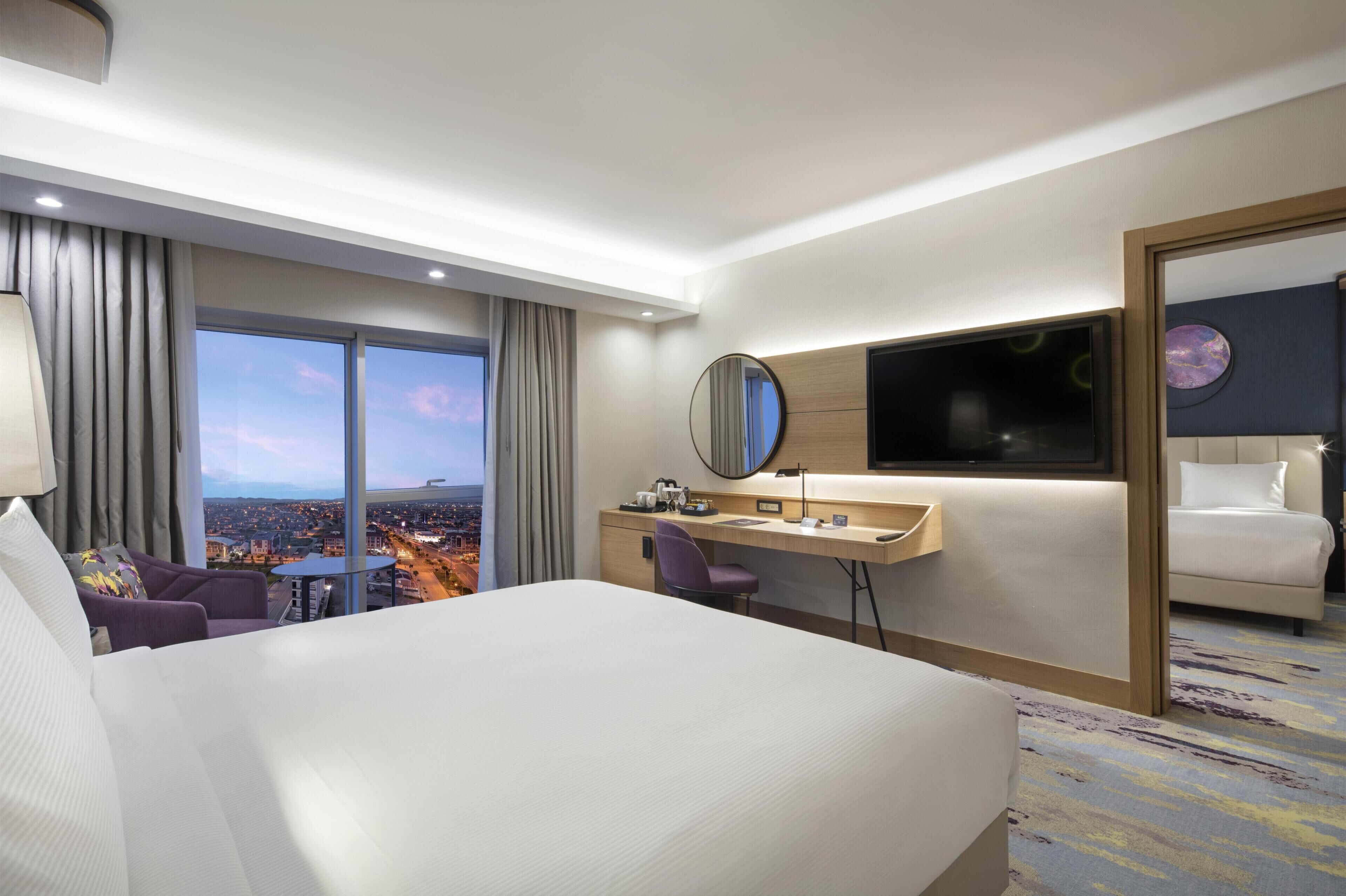 A hotel room with a large bed, a desk, a chair, a mirror, a TV, and a city view.