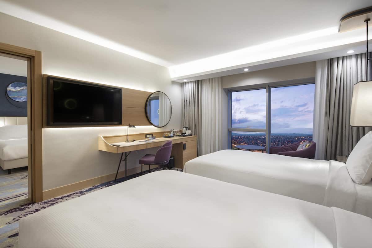 A hotel room with two beds, a desk, a chair, a TV, a large window with a city view, and a mirror.