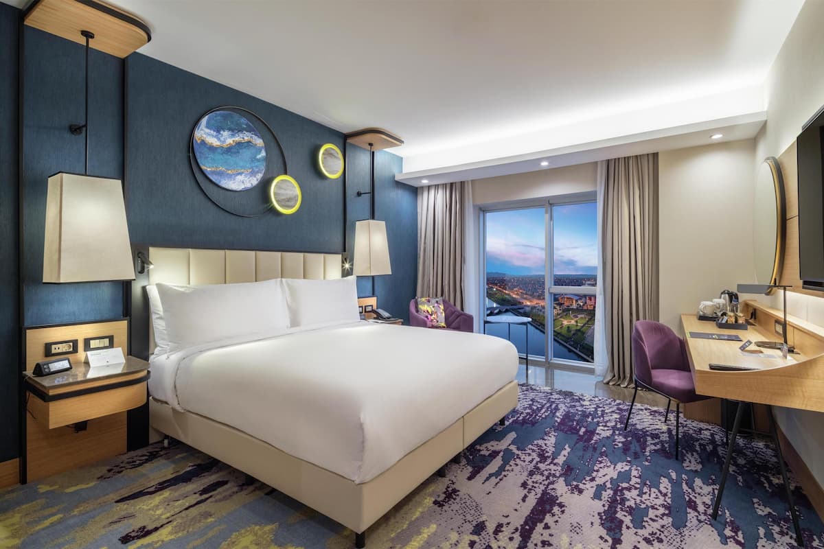 A modern hotel room with a large bed, a desk, a chair, a TV, and a view of the city.