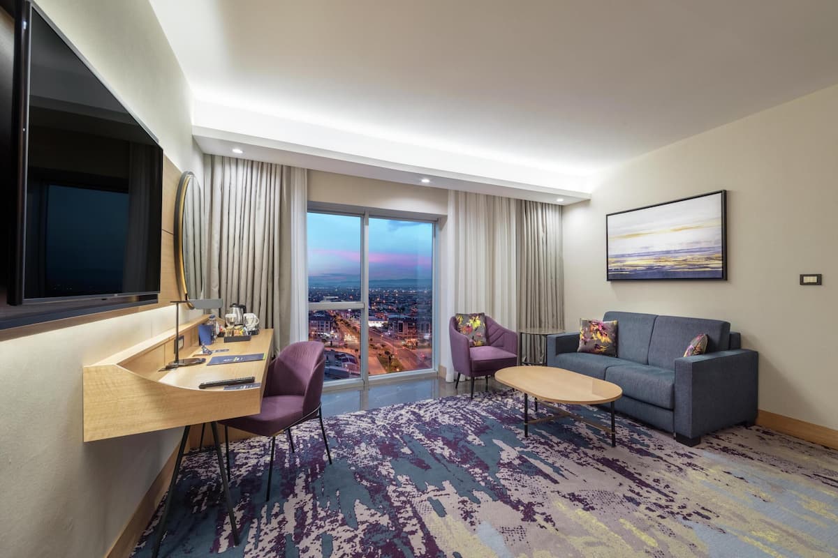 A hotel room with a city view, a sofa, a coffee table, a desk with a chair, and a large window with curtains.