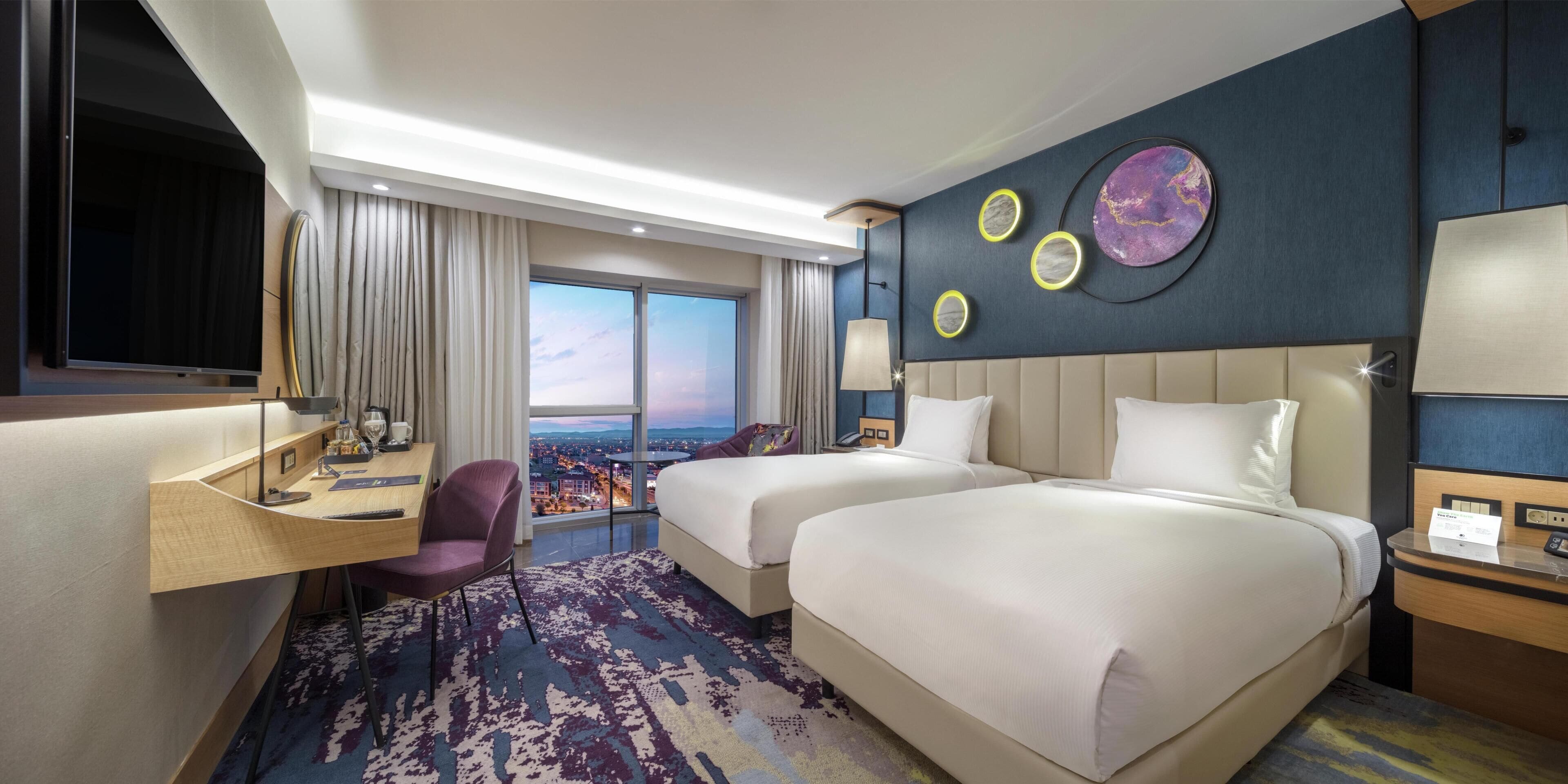 Une chambre d’hôtel avec deux lits, un bureau, une chaise, une télévision et une vue sur la ville.