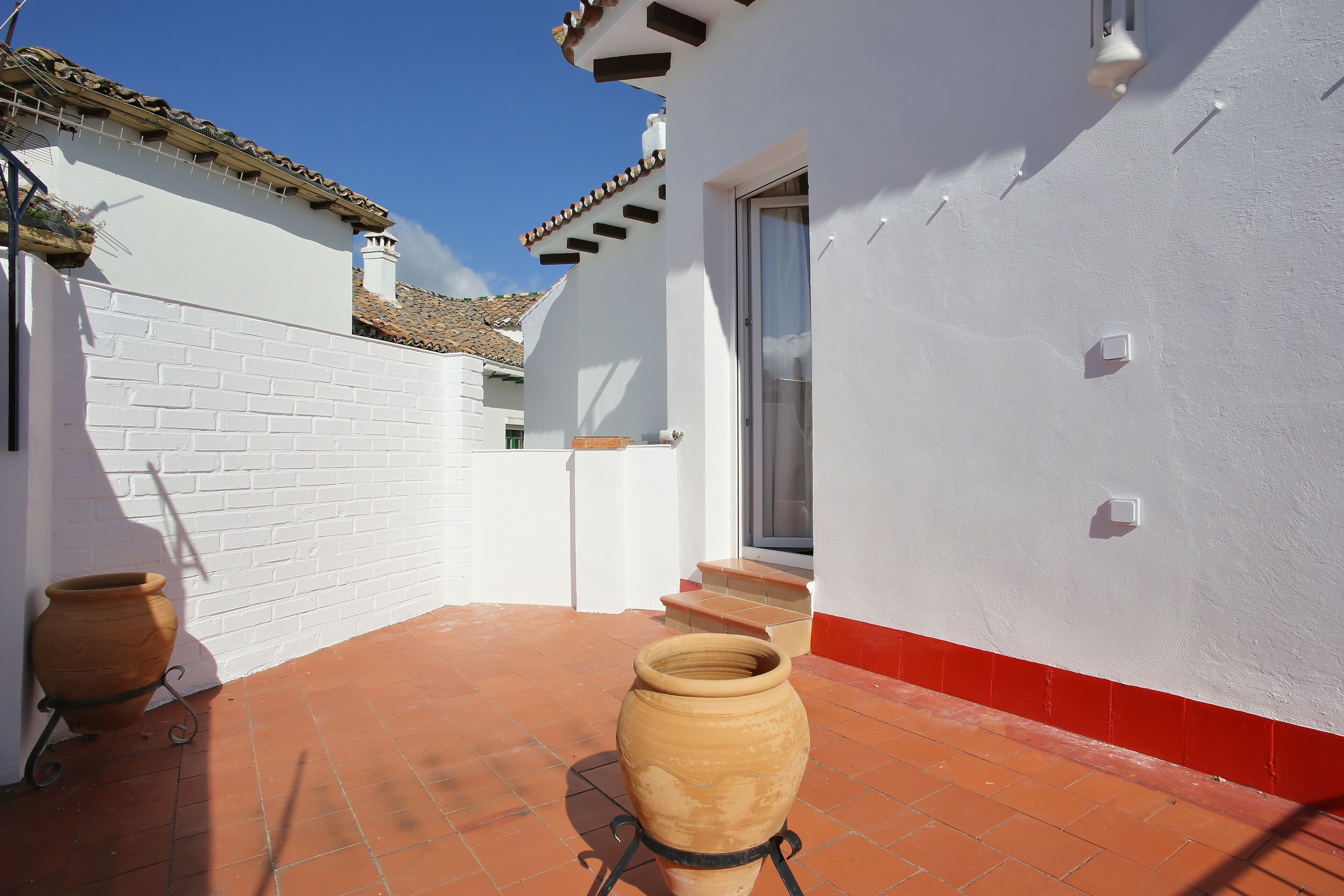 A white building with a red base, a tiled patio, and a large terracotta pot.
