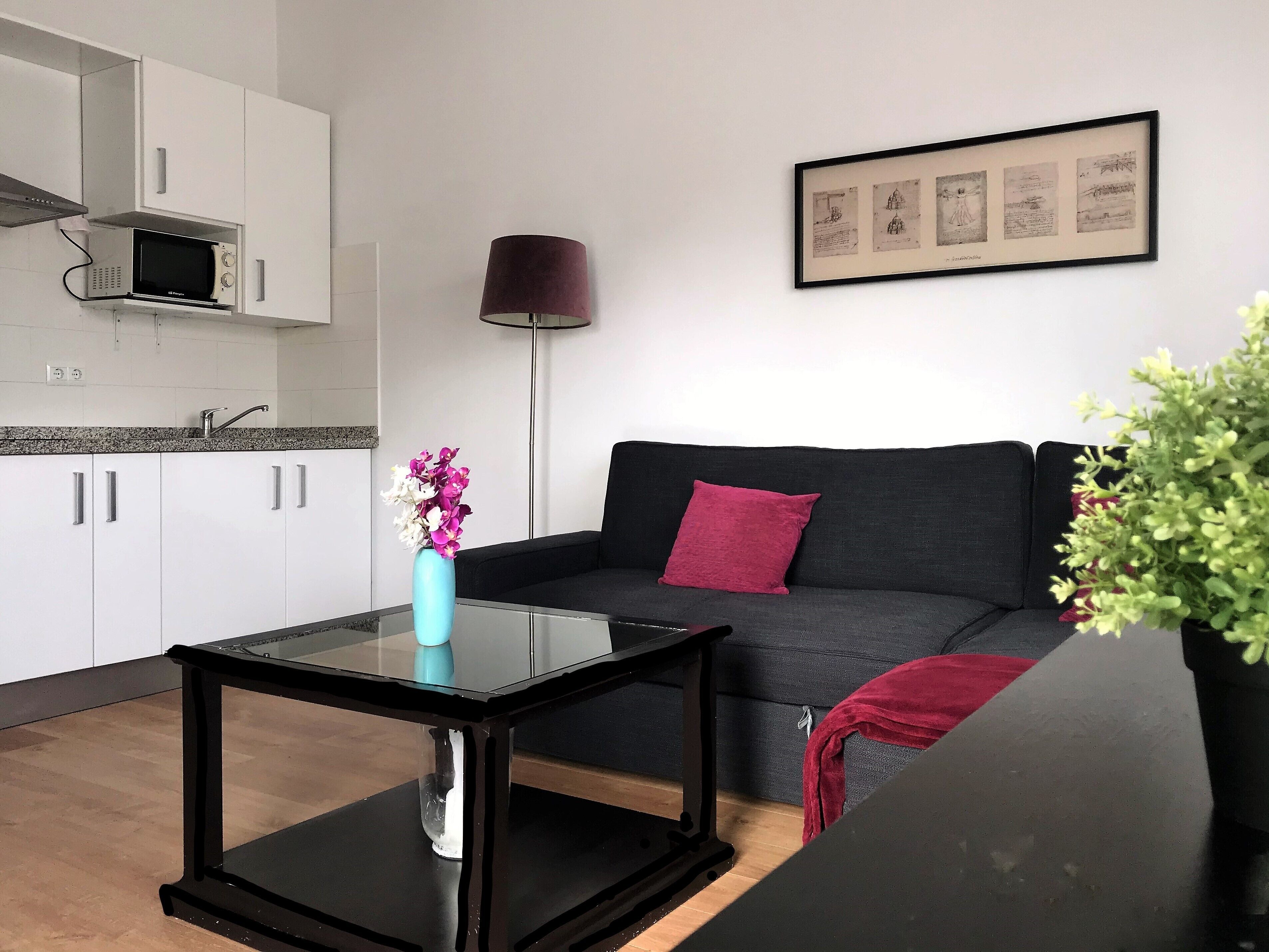 A modern living room with a glass coffee table, a dark grey sofa with a red throw, a black lamp, and a framed picture on the wall.