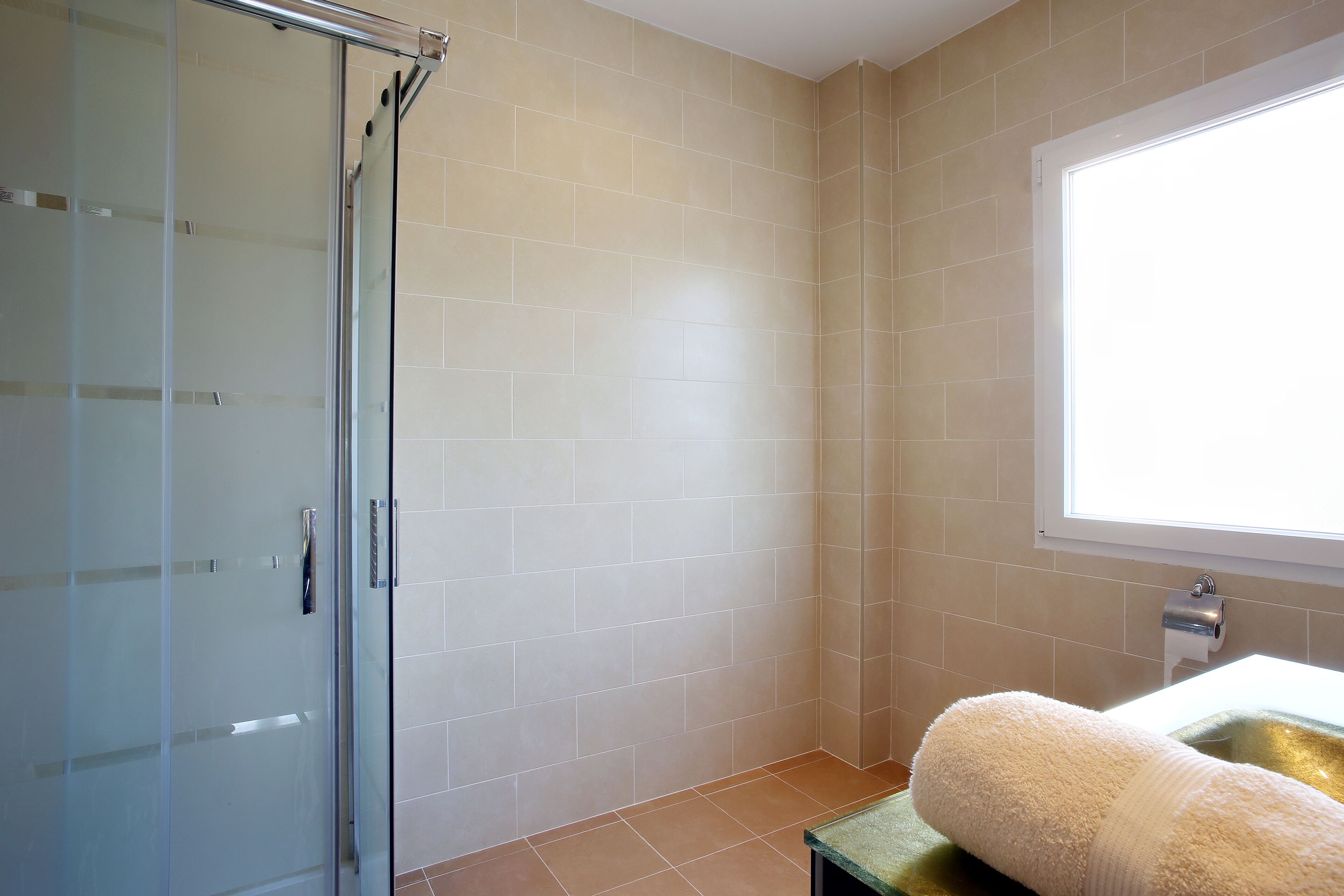 Un baño con cabina de ducha de vidrio, baldosas color crema, una ventana y una toalla sobre un tapete verde.