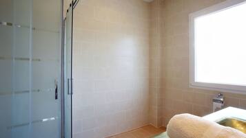 Un baño con cabina de ducha de vidrio, baldosas color crema, una ventana y una toalla sobre un tapete verde.
