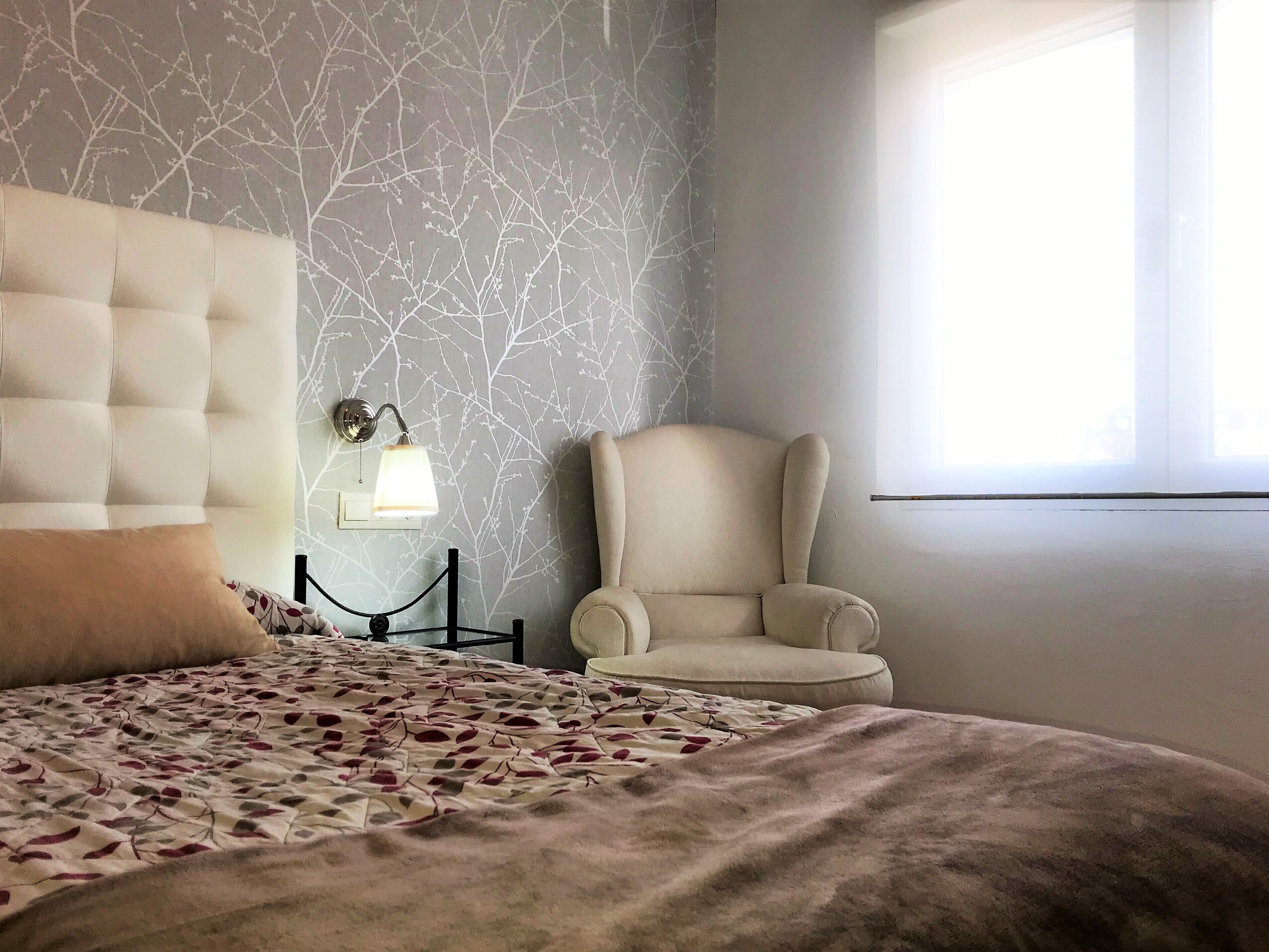 A bedroom with a tufted headboard, a large bed with a patterned bedspread, a bedside lamp, a chair, and a window with natural light.