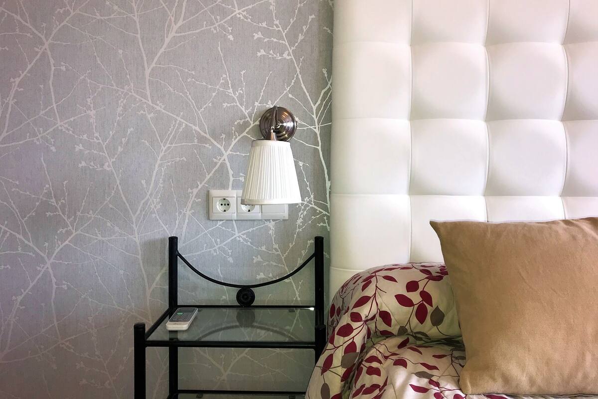 Une chambre à coucher avec un mur décoré d’un motif imitant le marbre, une tête de lit blanche à motifs capitonnés, une table de chevet avec une lampe et une couvre-lit à motifs.