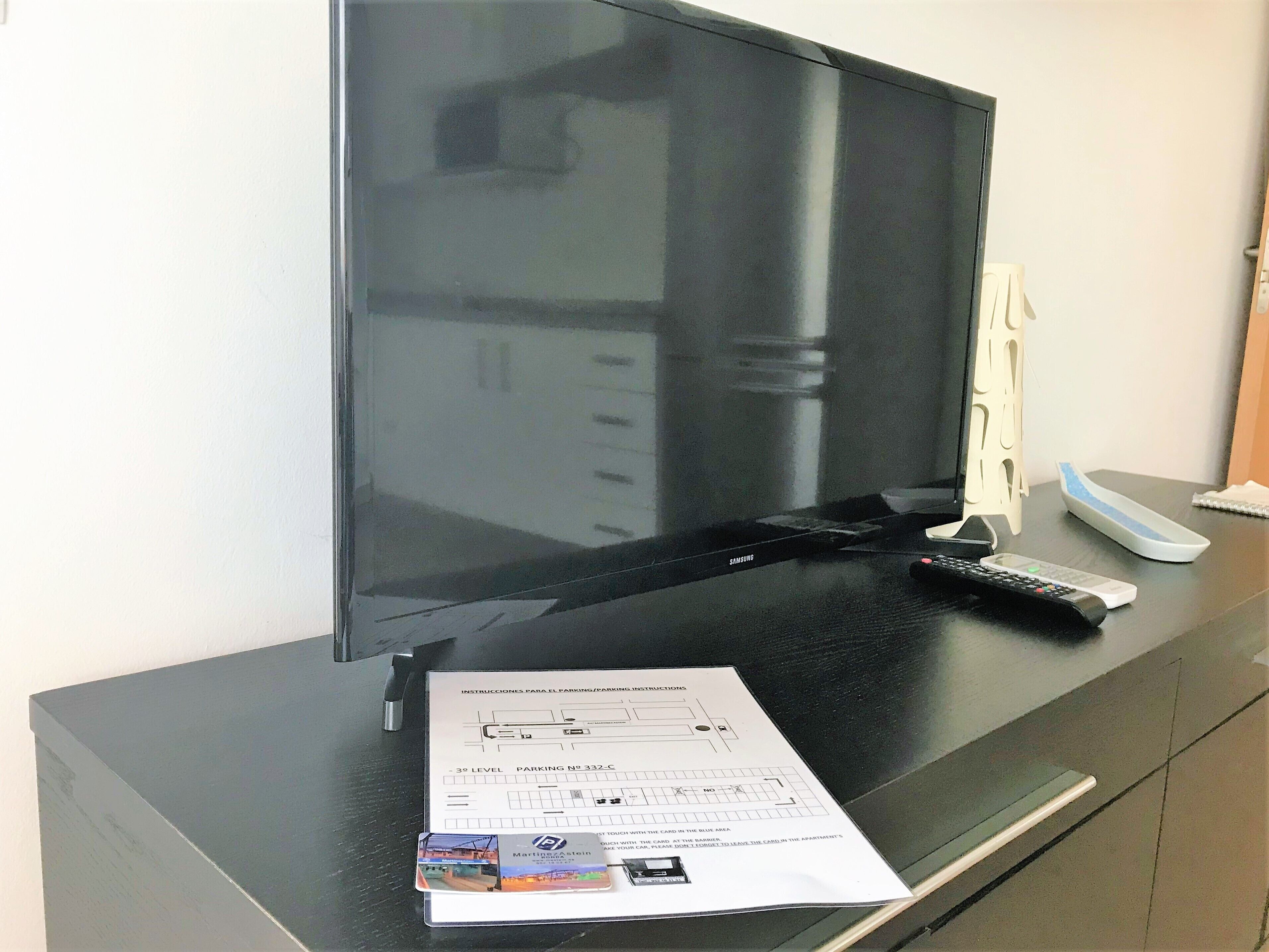 A flat-screen TV on a black TV stand, a remote control, a parking instructions paper, and a card on the stand.