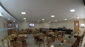 A dining area with multiple wooden tables and chairs, a television mounted on the wall, and a kitchen area in the background. - Hotel Portal da Amazônia (Varzea Grande)
