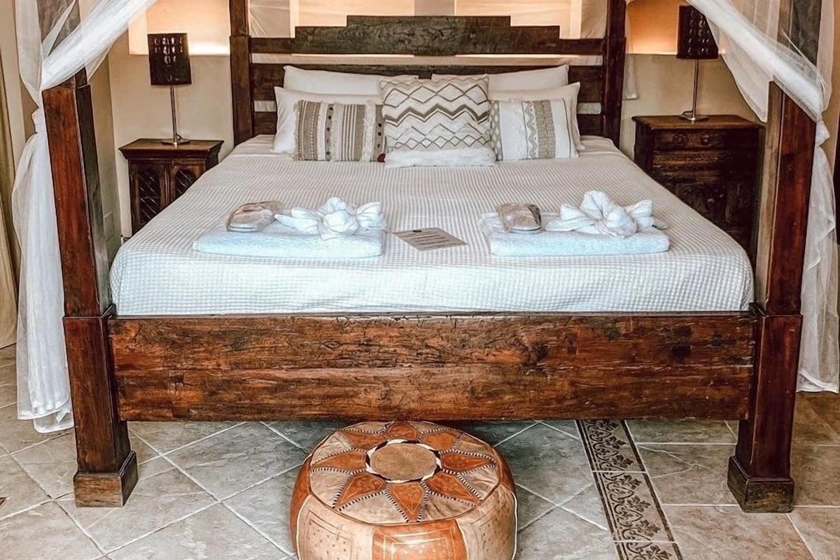 A four-poster bed with a wooden frame, white linens, and decorative pillows. There are two bedside tables with lamps. A round ottoman is placed in front of the bed.