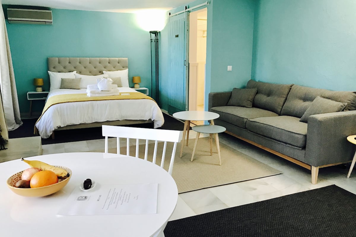 A hotel room with a bed, a sofa, a small table with a bowl of fruit, and a white chair.