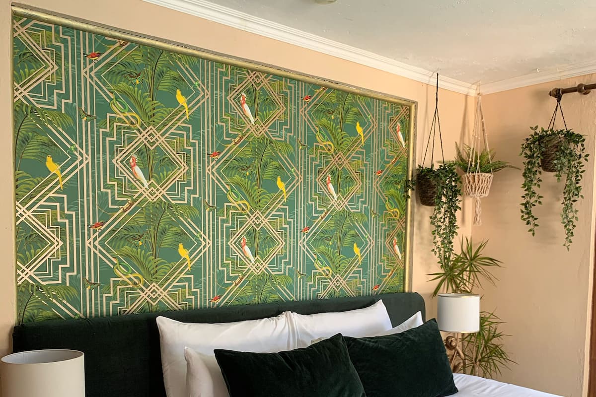 A bedroom with a green patterned headboard, a bed with white and dark green bedding, two bedside lamps, and hanging plants.
