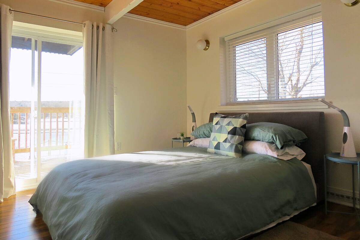 A bedroom with a bed, a nightstand, a window with blinds, and a view of a balcony.