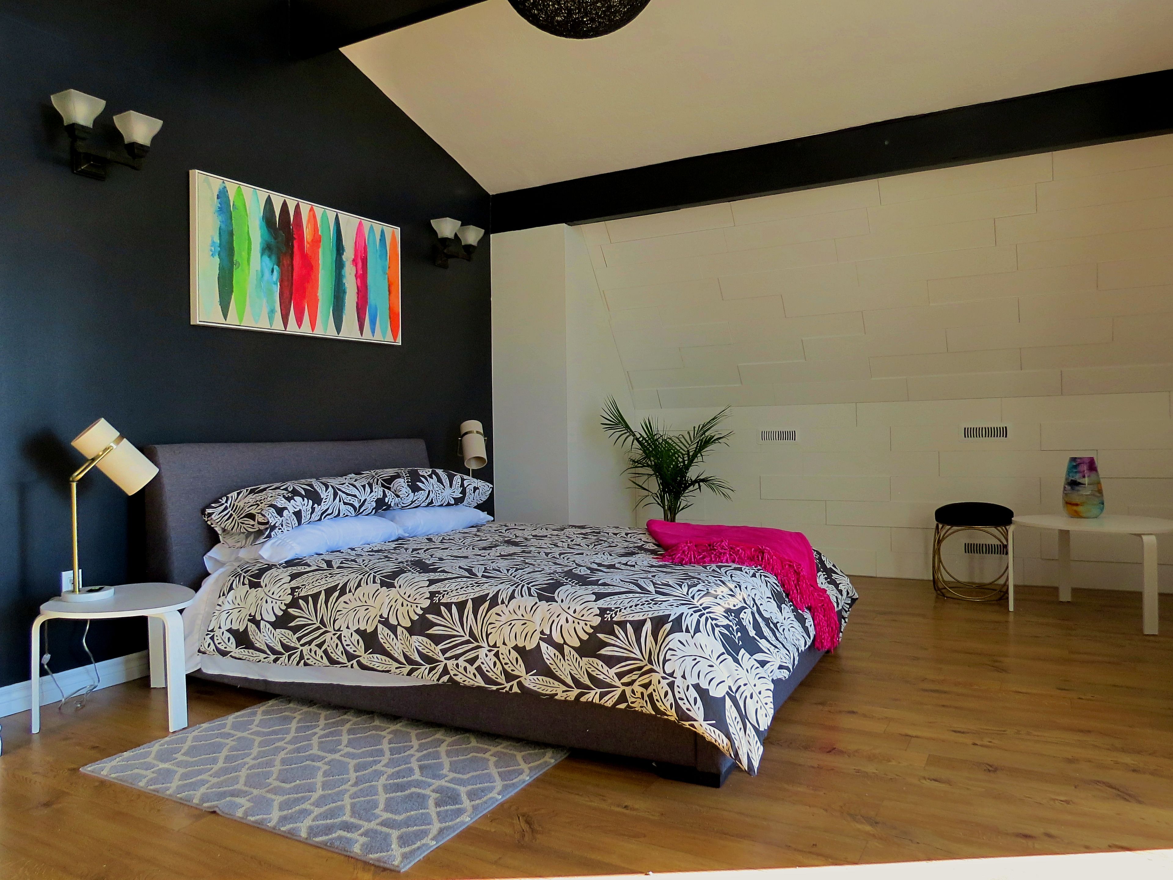 A modern bedroom with a bed, bedside tables, a floor lamp, a plant, and a colorful abstract painting on the wall.