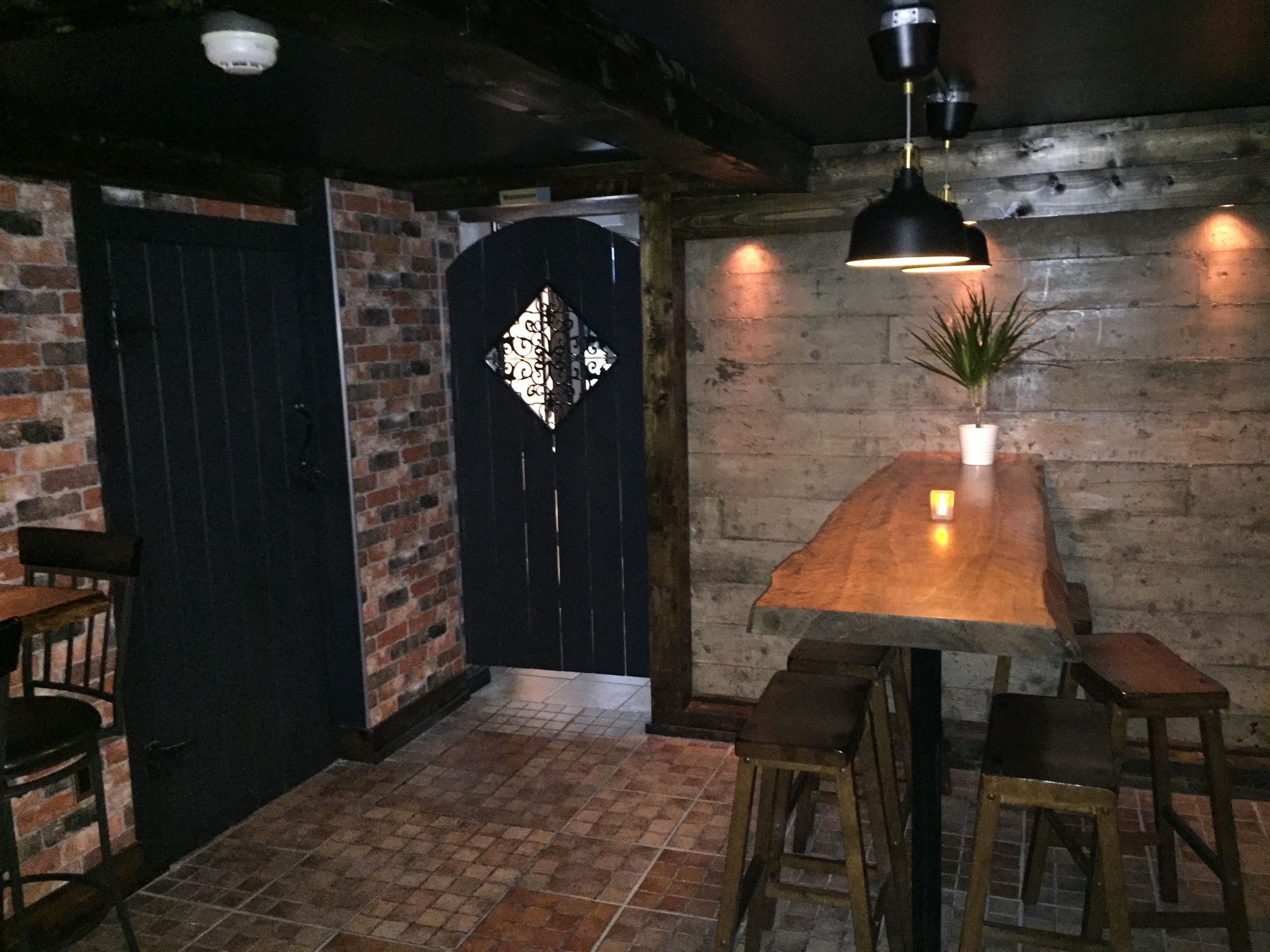 A rustic interior with a wooden table, stools, a brick wall, and a black door with a decorative pattern.