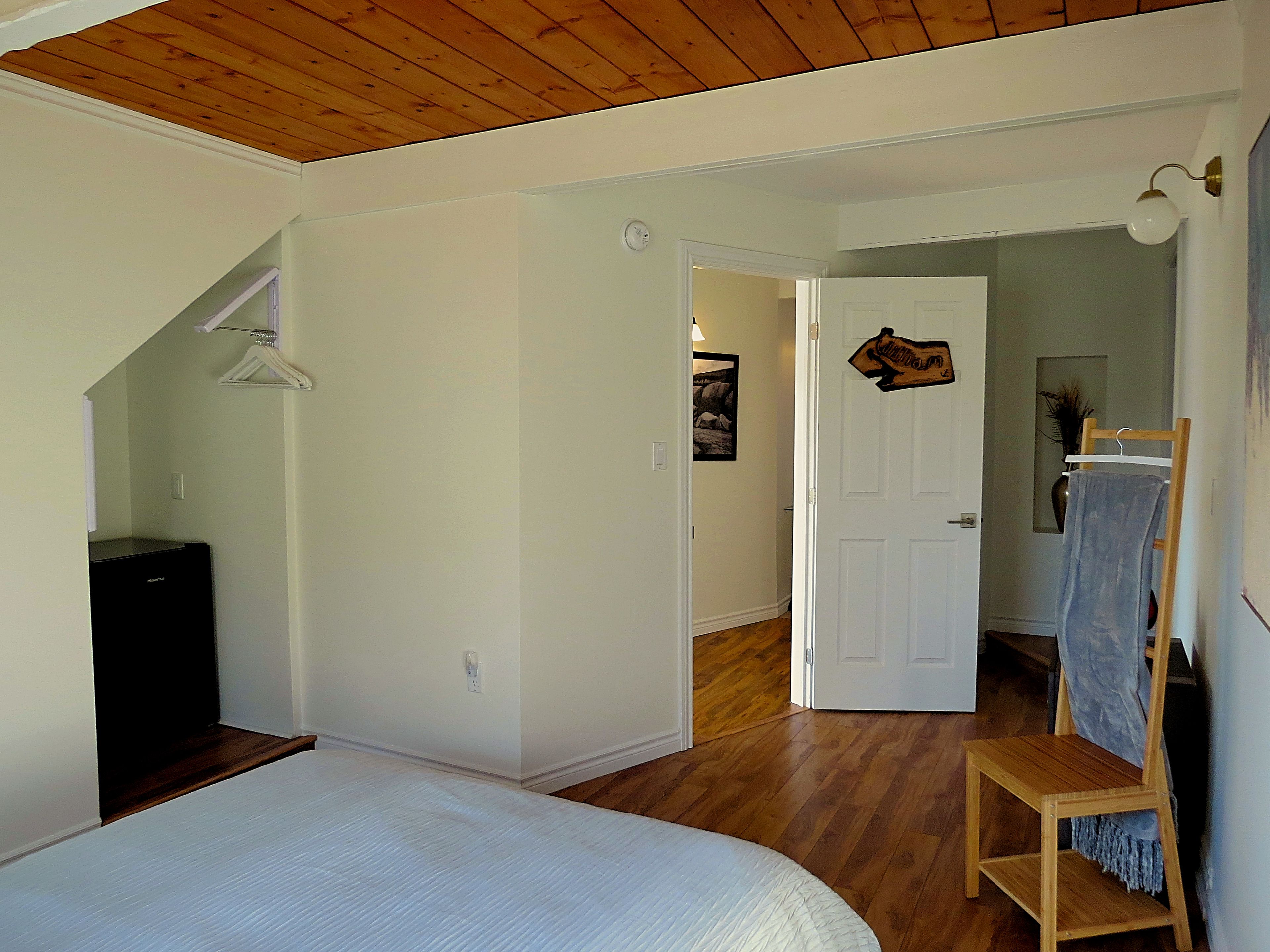 Une chambre à coucher avec un lit, un plancher en bois, une porte blanche et un couloir menant à une autre pièce.