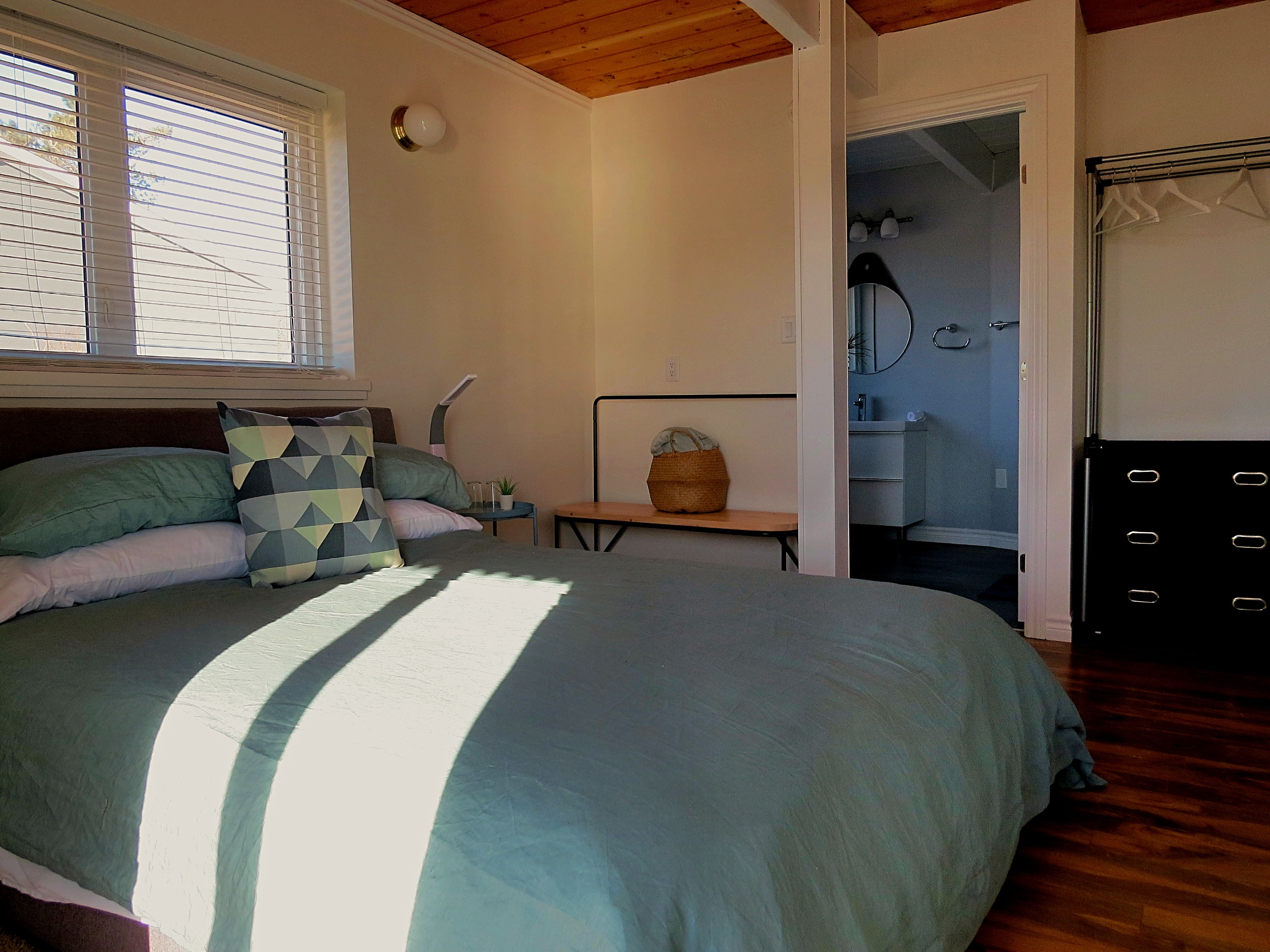Une chambre avec un grand lit, une table de chevet, une lampe, une petite plante et un panier. Une porte donne accès à une salle de bain équipée d’un miroir et d’un porte-serviettes.
