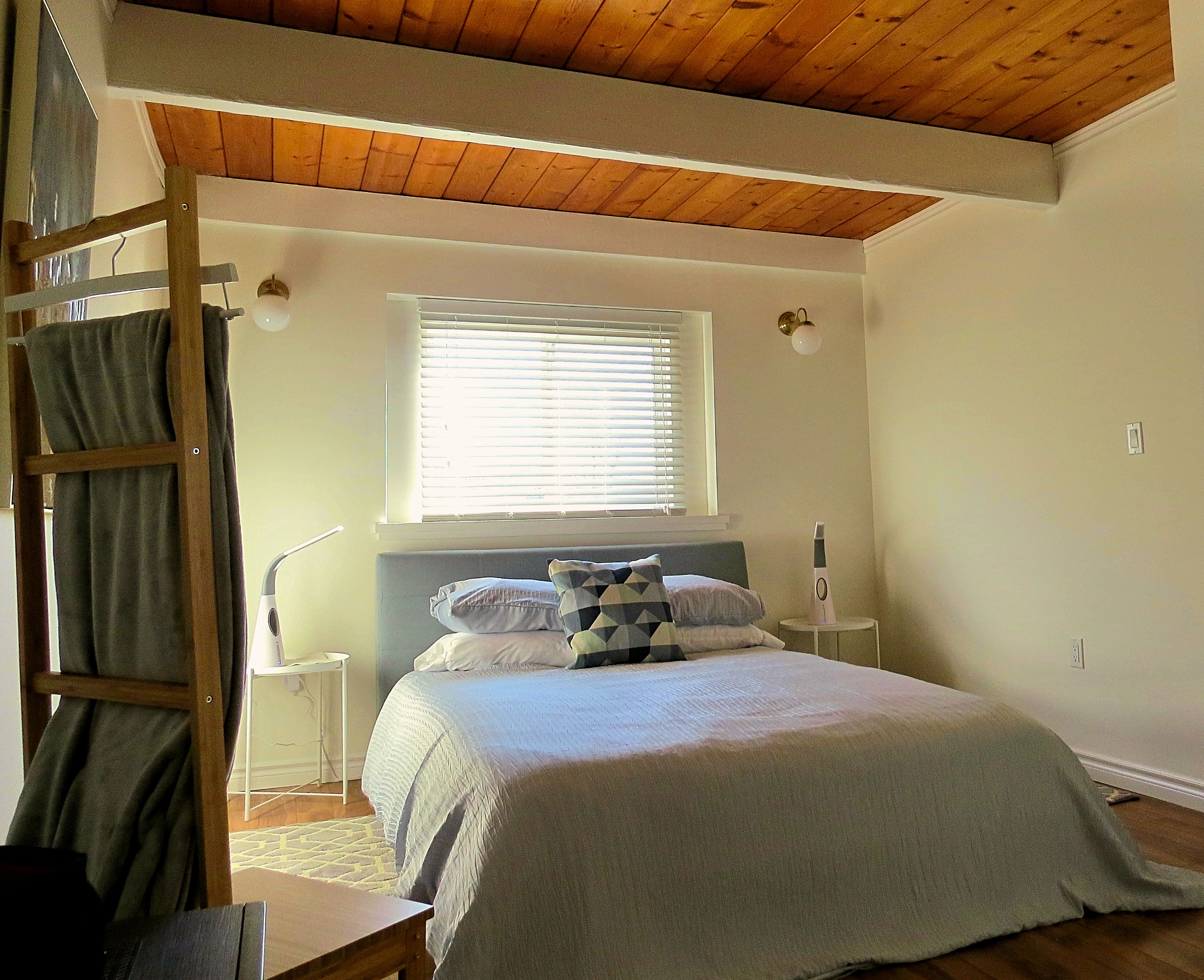 Une chambre avec un plafond en bois, un lit recouvert d’une couverture blanche et grise, une fenêtre avec des stores et une table de chevet avec une lampe.