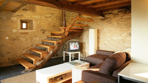 A rustic stone-walled room with a wooden staircase, a white sofa, and a television on a small table. - A parada das bestas (Palas de Rei)