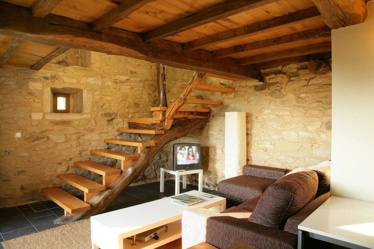 Ein rustikaler Raum mit Steinwänden, einer Holztreppe, einem weißen Sofa und einem Fernseher auf einem kleinen Tisch.
