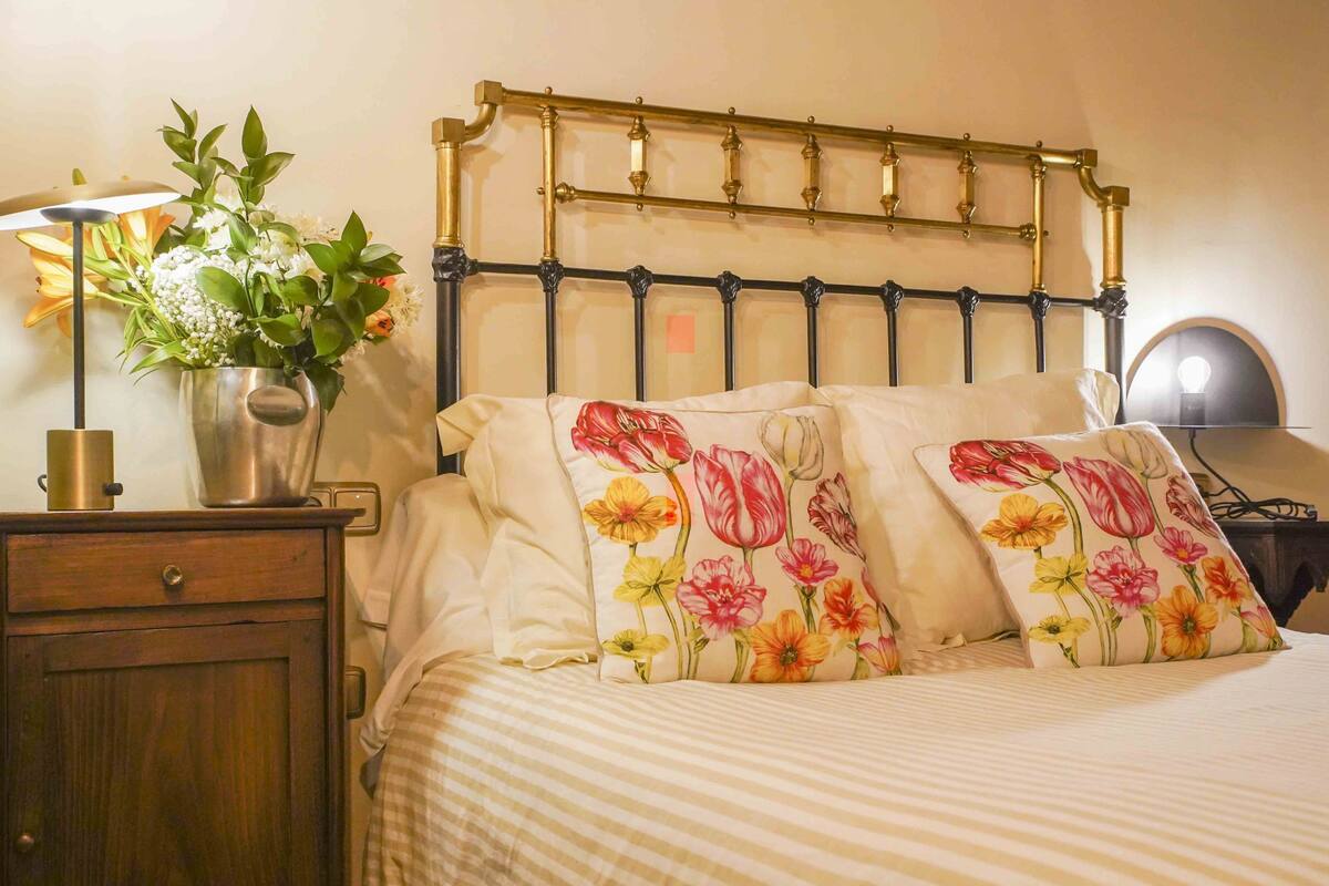 Ein ordentlich bezogenes Bett mit gestreifter Bettwäsche, einem Metall-Kopfstück und blumigen Kissen.