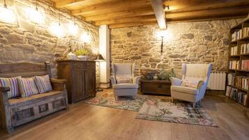 A cozy living room with a stone wall, wooden ceiling beams, a sofa, two armchairs, a coffee table, a bookshelf, and a radiator.