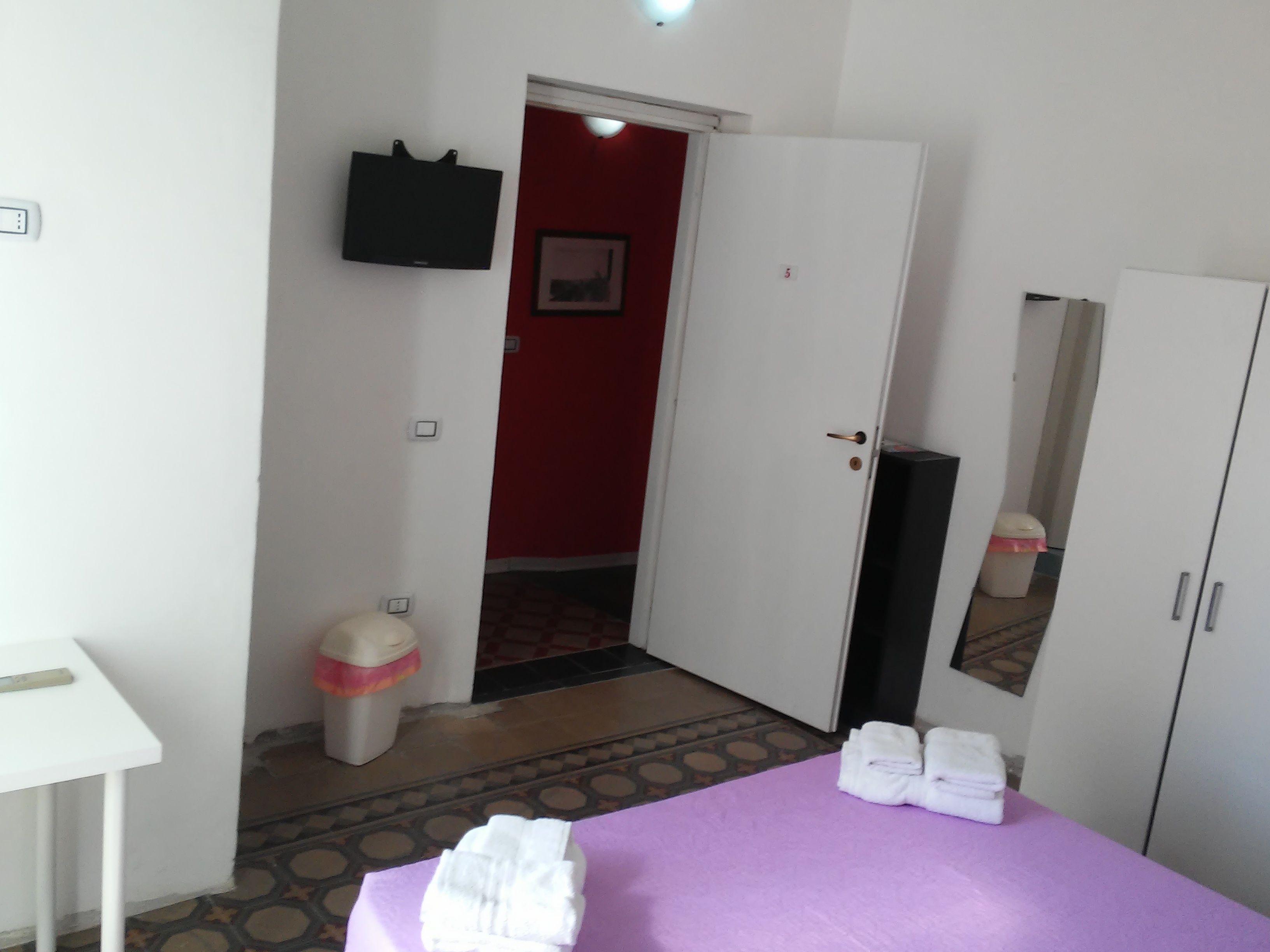 Une chambre d’hôtel avec un lit, une armoire blanche, une petite table, une poubelle et un téléviseur fixé au mur.