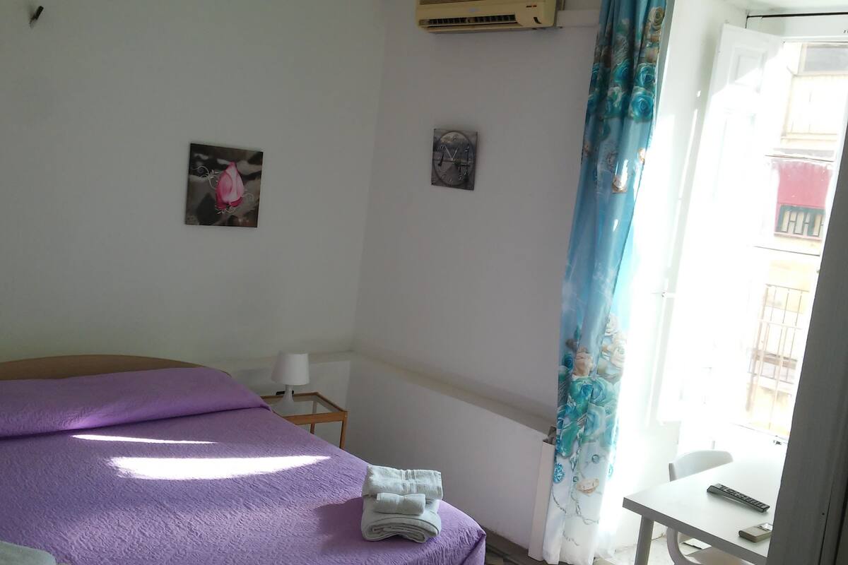 Ein Schlafzimmer mit einem Bett, einem kleinen Tisch, einem Sessel, einer Klimaanlage, einer Uhr und einem Fenster mit Vorhang.