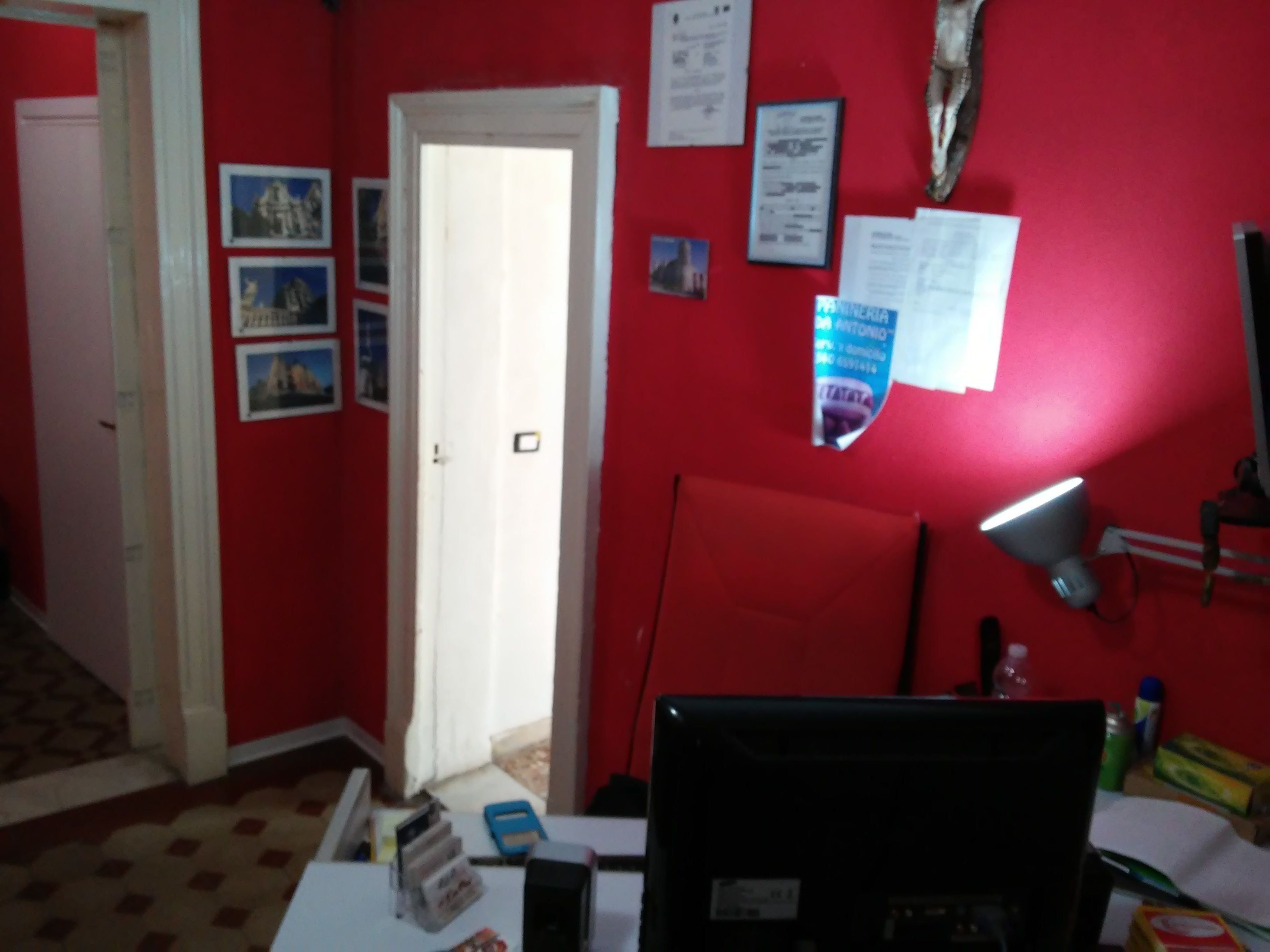 A room with a red wall, a white door, and a desk with a computer monitor. There are papers and a lamp on the wall.