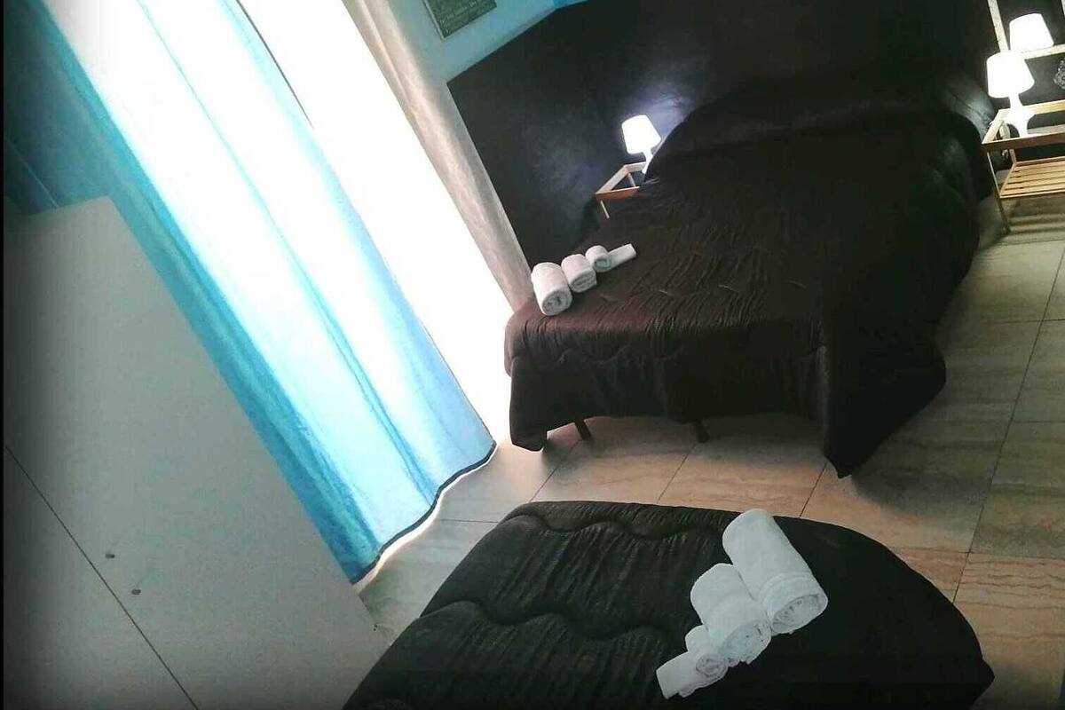 Ein Zimmer mit einem Bett, einem Sessel und einem kleinen Tisch. Es gibt ein Fenster mit Vorhängen, eine Wandklimaanlage und einen Spiegel.