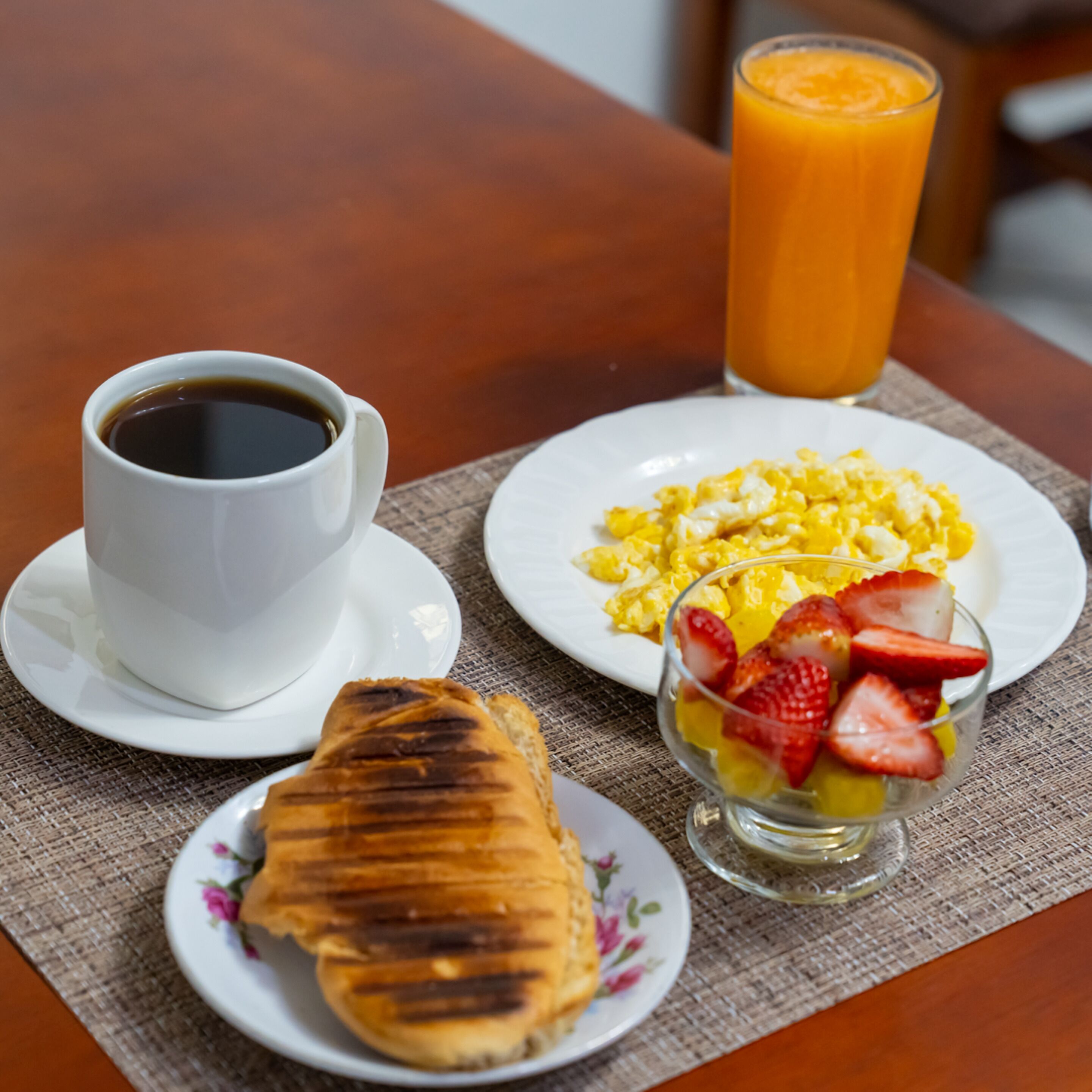 Un desayuno con una taza de café, huevos revueltos, un croissant y un vaso de jugo de naranja sobre una mesa.