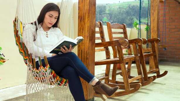 Una persona sentada en una hamaca leyendo un libro, con una mecedora de madera y una vista de paisaje verde al fondo.