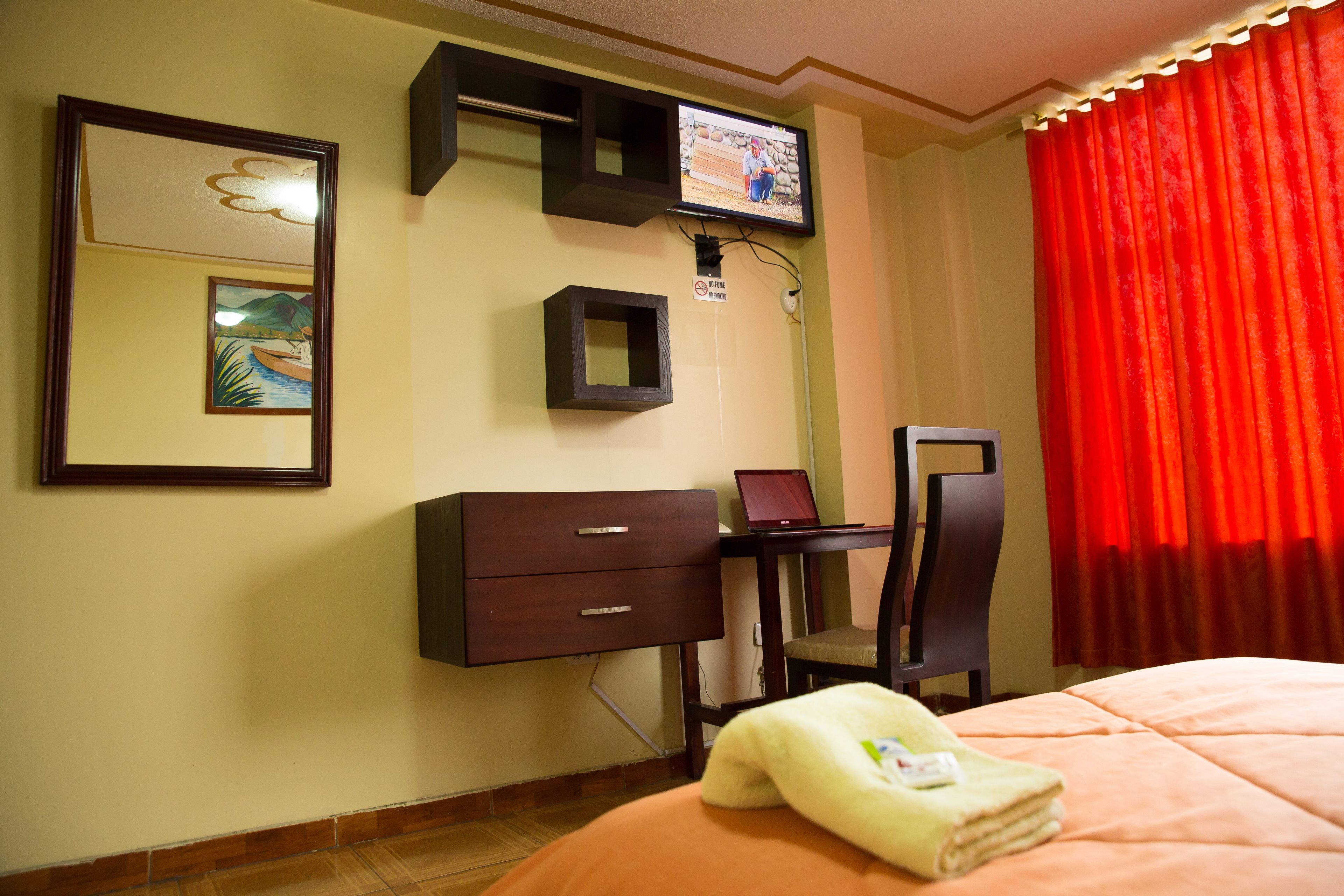 Ett hotellrum med en säng, ett skrivbord med en bärbar dator, en stol, en tv som är monterad på väggen och en tavla på väggen.