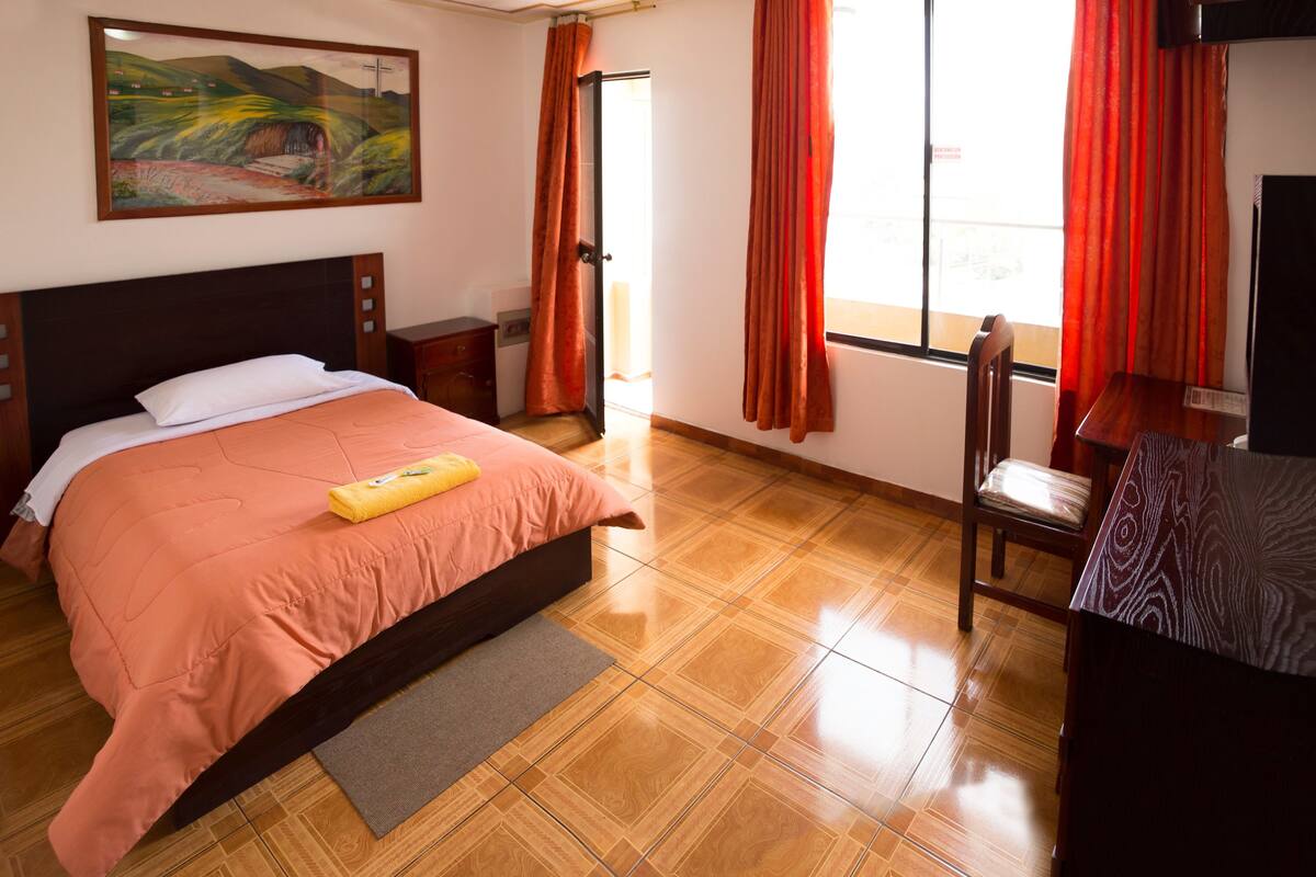 Ein Hotelzimmer mit Bett, Sessel, Schreibtisch, einem Bild an der Wand und einem Fenster mit Vorhängen.