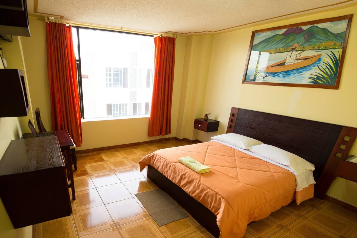 Ein Hotelzimmer mit Bett, Schreibtisch, Stuhl, einem Fenster mit Vorhängen, einem Bild an der Wand und einem kleinen Nachttisch.