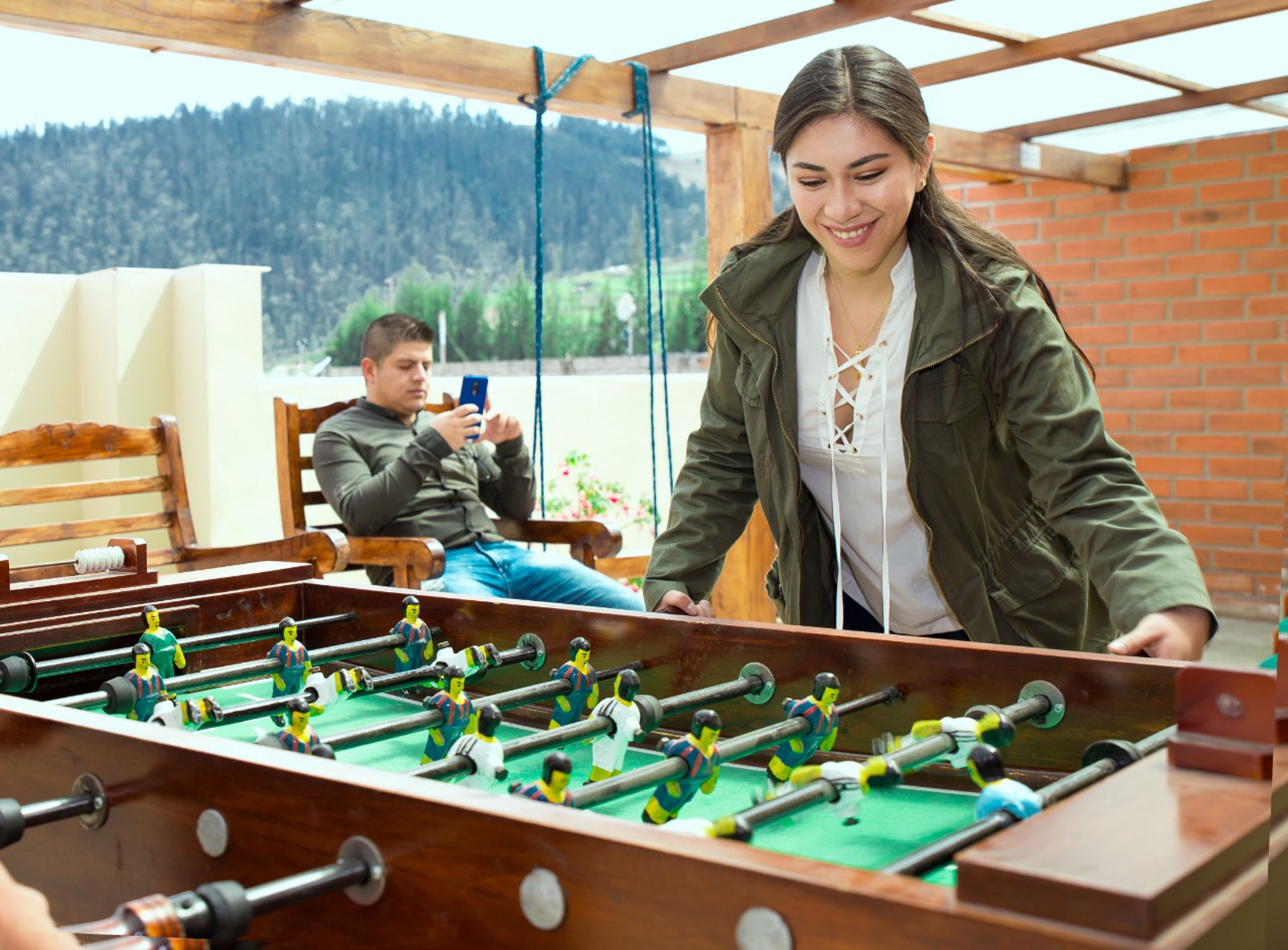 A wooden foosball table in a room with a brick wall and a view of a forested area.