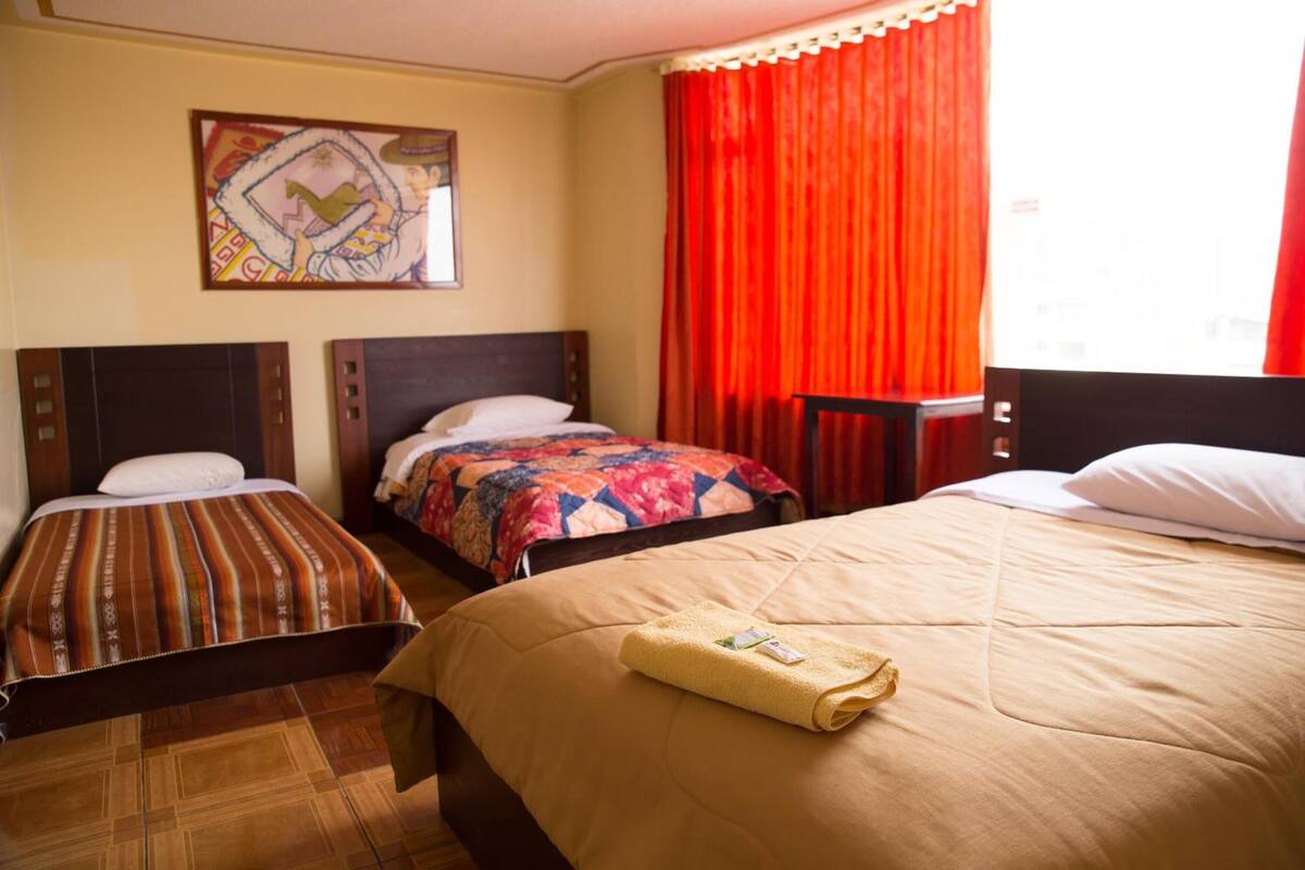 Ein Hotelzimmer mit zwei Einzelbetten, einem großen Bett, einem Fenster mit roten Vorhängen und einem Bild an der Wand.