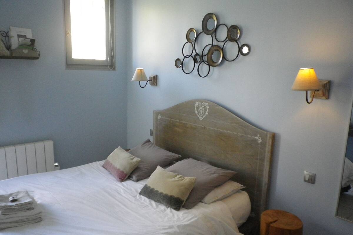 A neatly made bed with white linens and multiple pillows, a wooden headboard with a heart design, two wall-mounted lamps, and a radiator on the wall.