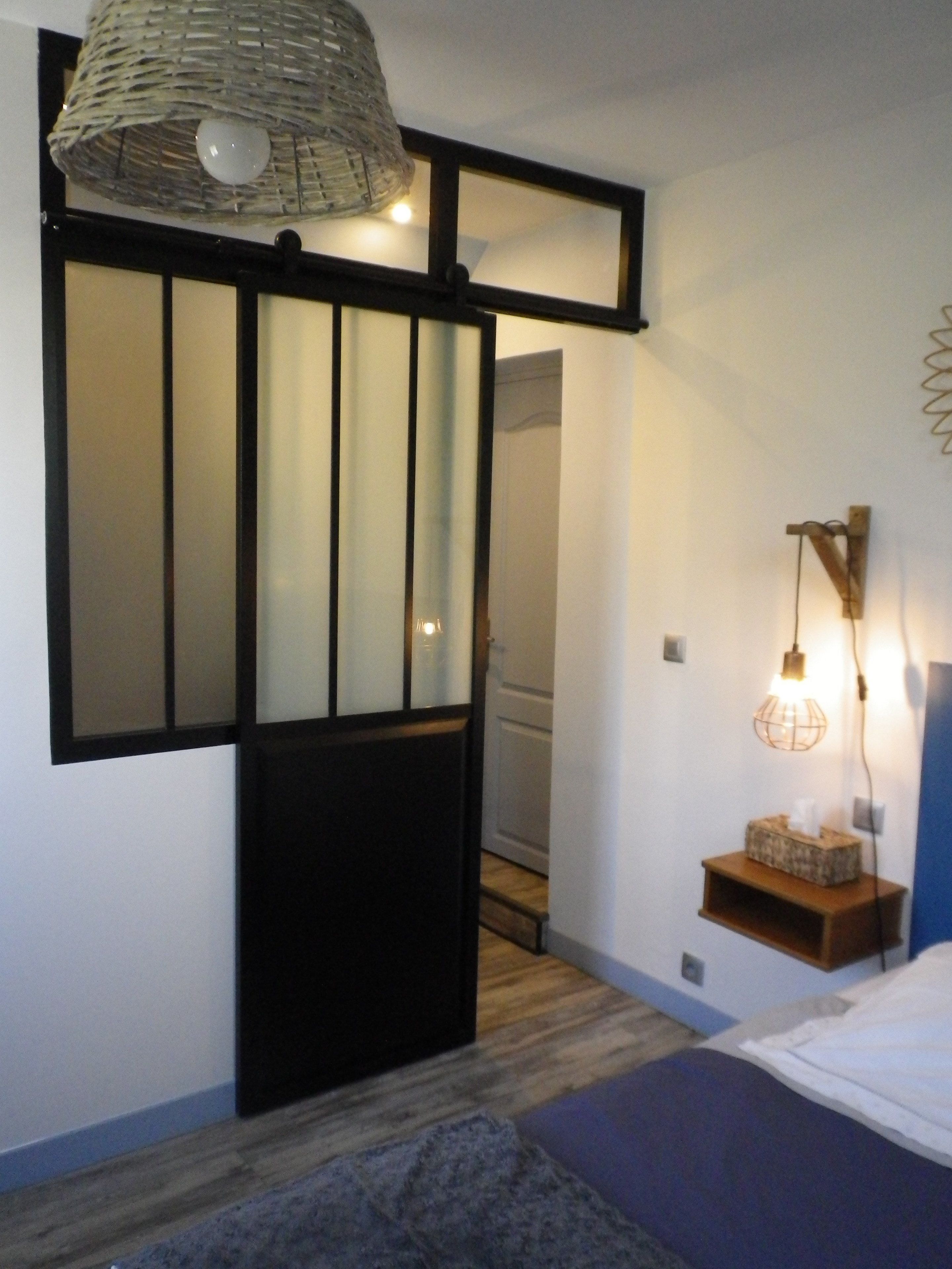 Een slaapkamer met een bed, een nachtkastje met een lamp, een wandplank en een geweven lamp.