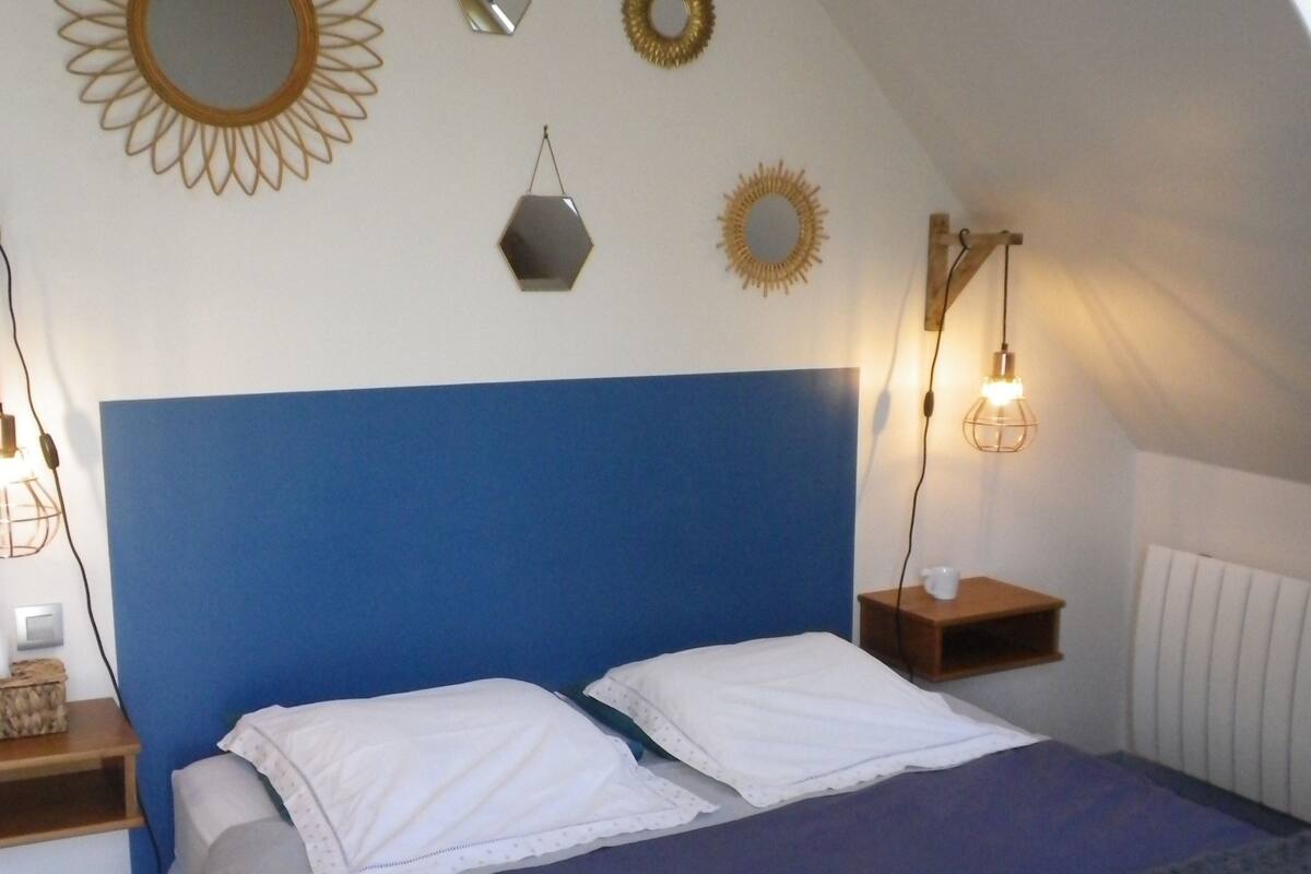 A bedroom with a blue headboard, two white pillows, a bed with a dark blue blanket, and a bedside table with a lamp and a cup.