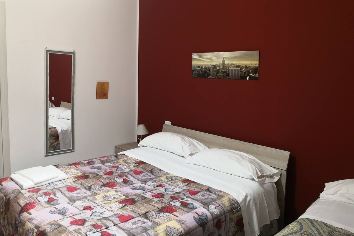 Une chambre d’hôtel avec un lit, une table de chevet, un miroir, un tableau représentant un paysage urbain et un mur d’accent rouge.