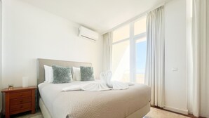 A neatly made bed with white linens and two green pillows, a wooden nightstand with a drawer, and a large window with curtains. - Flat Praia Rocha Balconies Lotus (Portimão)