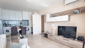 A modern living room with a flat-screen TV, a sofa, and a coffee table. There is a kitchen area with cabinets and a dining table. - Flat Praia Rocha Balconies Lotus (Portimão)
