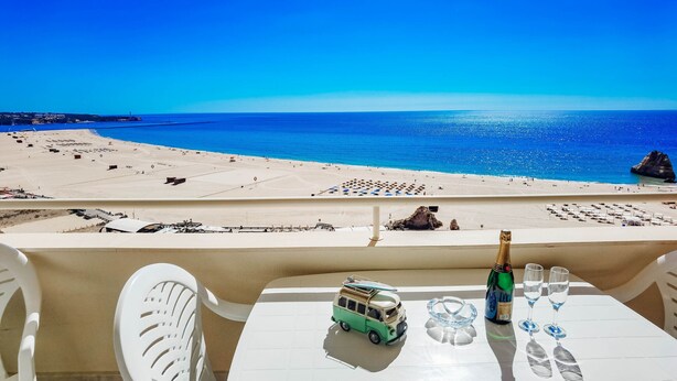 A balcony with a table set for two, overlooking a beach with clear blue water and a sandy shore. - Flat Praia Rocha Balconies Lotus (Portimão)
