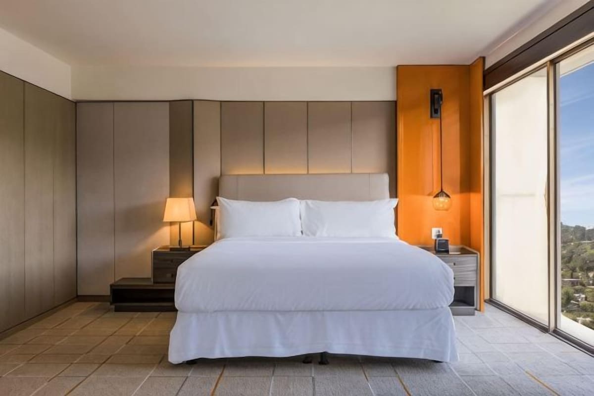 A hotel room with a large bed, a nightstand, a lamp, and a view through a large window.