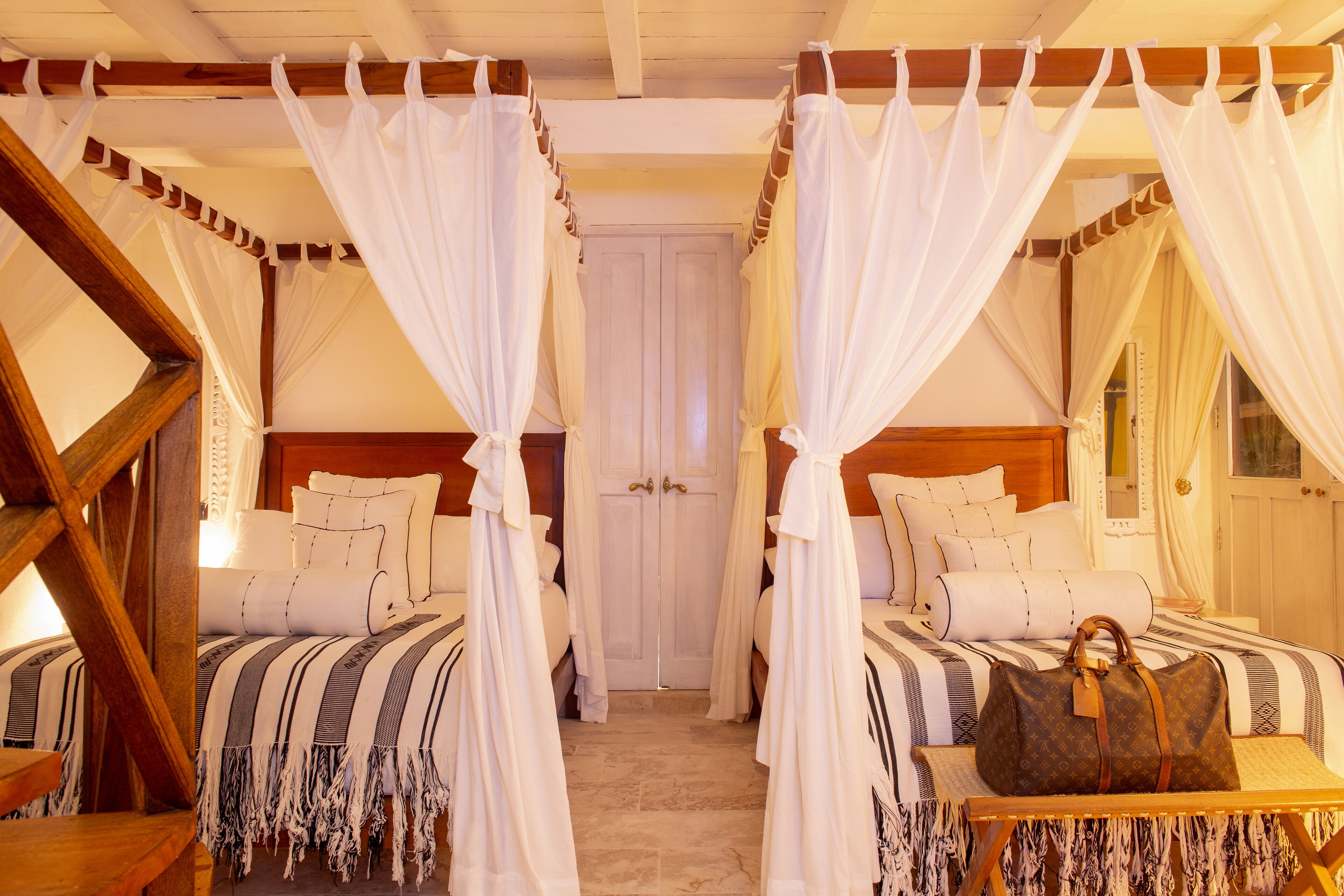 A room with two beds, each with white bedding and a canopy. There is a wooden table with a bag on it.